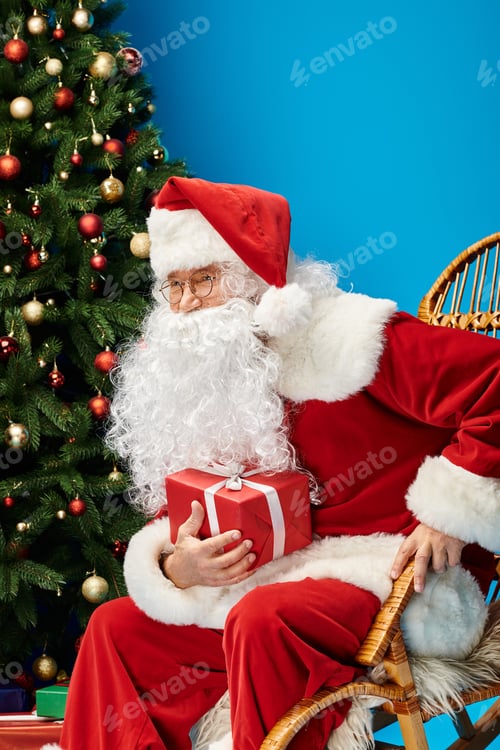 Preview: happy Santa Claus with beard and eyeglasses sitting in rocking chair with gift near Christmas tree