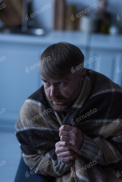 Preview: displeased and frozen man looking away while sitting under warm blanket during power blackout