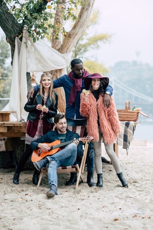 Preview: Happy young people with cocktails at alfresco picnic enjoying guitar on sandy beach