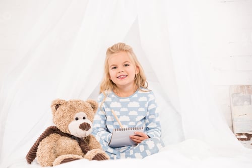 Preview: adorable child in pajamas sitting on bed with notebook and teddy bear