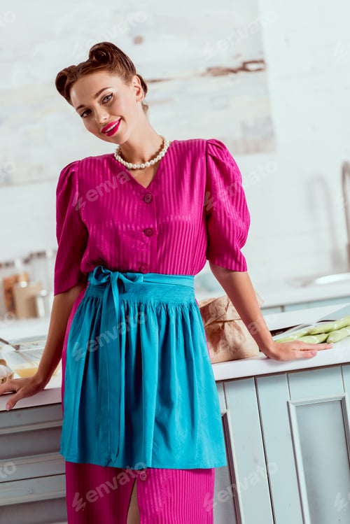 Preview: Elegant pin up girl in crimson dress and blue apron standing by kitchen table