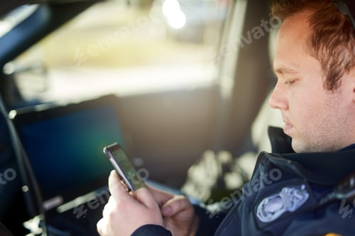 Preview: Cropped shot of a handsome young male police officer using his cellphone while out on patrol