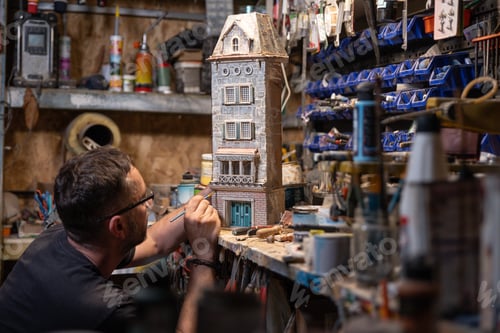 Preview: Modeller man working at scale model of miniature building in his workshop full of tools