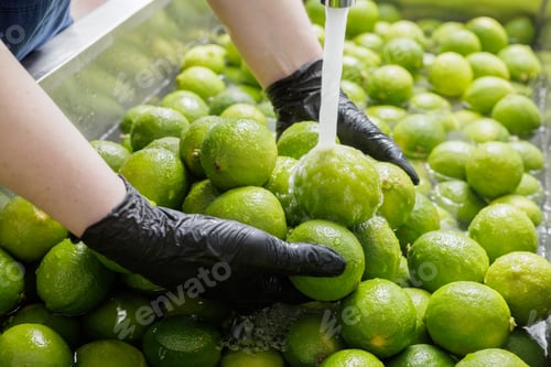 Preview: Gloved hands washing green limes under water stream at fruit cleaning station.