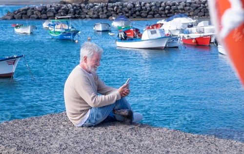 Preview: Senior bearded man in excursion at sea, sitting at harbour using mobile phone. Horizon over water