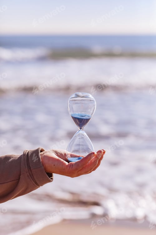 Preview: Time is running fast. Hourglass with blue sand inside in mature woman's hand symbolize the brev