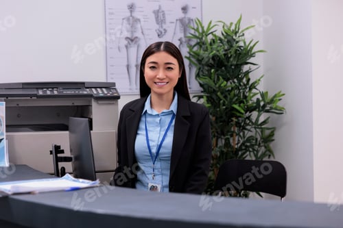 Preview: Smiling asian receptionist standing at hospital reception counter scheduling follow-up appointments