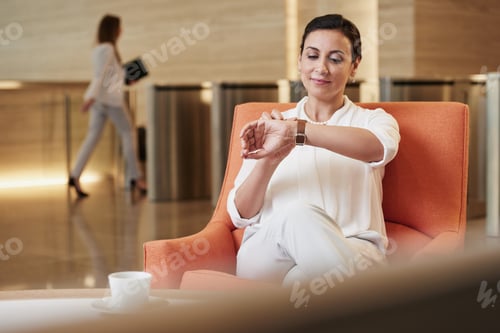 Preview: Business woman checking her watch while sitting in airport waiting lounge for business travel