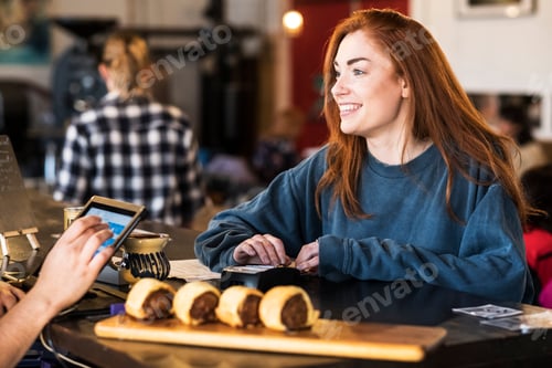 Preview: Smiling young woman with long red hair standing at a counter in a restaurant, paying her bill.