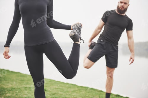 Preview: Fitness couple stretching outdoors in park near the water. Young bearded man and woman exercising
