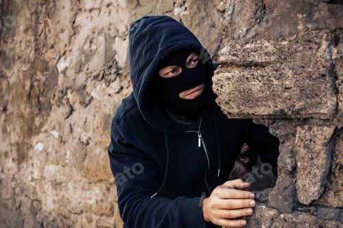 Preview: Man in mask behind stone wall watching someone outdoors. Criminal activity