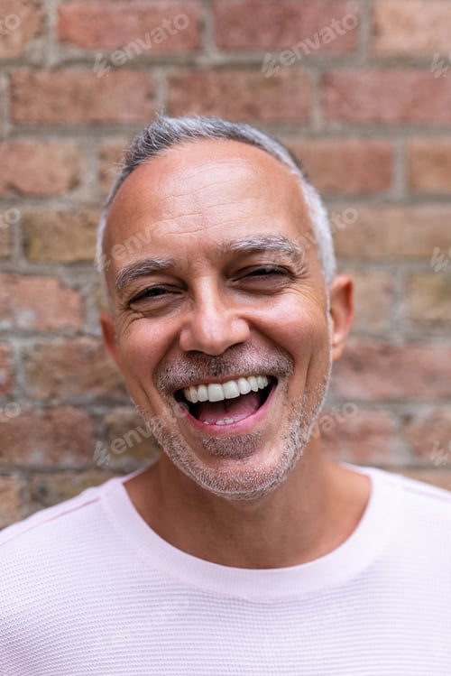 Preview: Adult man wearing pink sweater laughing looking at camera. Orange brick wall background. Vertical.