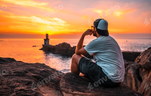 Preview: A young man sitting in a white shirt and cap looking at the orange sunset