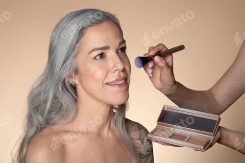 Preview: Makeup artist holding contouring palette and doing makeup to attractive senior woman, aged female