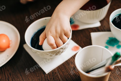 Preview: Easter day. Kid boy with dirty hands painting eggs on wooden background. Child sitting in a kitchen
