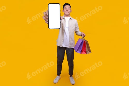 Preview: Asian Man Showing Blank Phone Screen While Bring Shopping Bags Isolated on Yellow Background