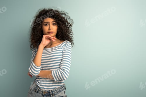 Vista previa: disappointed mixed race girl holding hand near chin while looking at camera on grey background