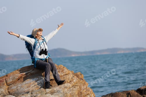 Preview: A senior woman sits on a rocky shoreline with arms outstretched, embracing the fresh sea breeze.