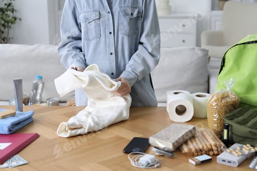 Preview: Survival kit. Woman packing different emergency supplies at wooden table indoors, closeup