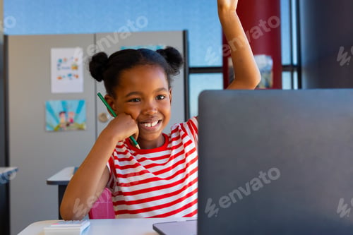 Preview: African american girl raising her hand while on video call on laptop in class at elementary school