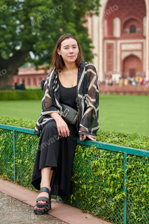 Preview: Attractive east asian woman in black dress sitting on fence in city park, girl outdoor resting