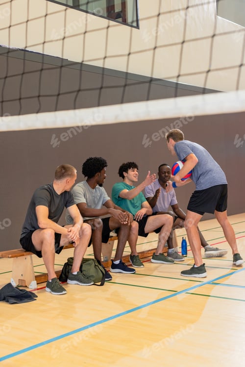 Preview: Diverse male volleyball partners sitting on bench and standing holding volleyball in school gym