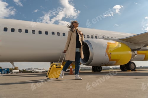 Preview: Passenger woman in sunglasses with luggage is outdoors near plane at airport looking away