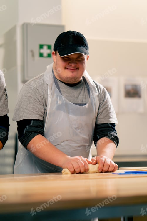 Preview: in a professional kitchen an inclusive worker in white uniform rolls out dough and smiles