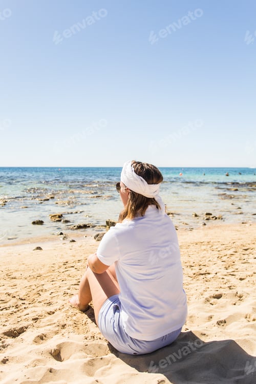 Preview: Holidays, summer, vacation, travel and people concept - Young woman sitting on the beach looking at