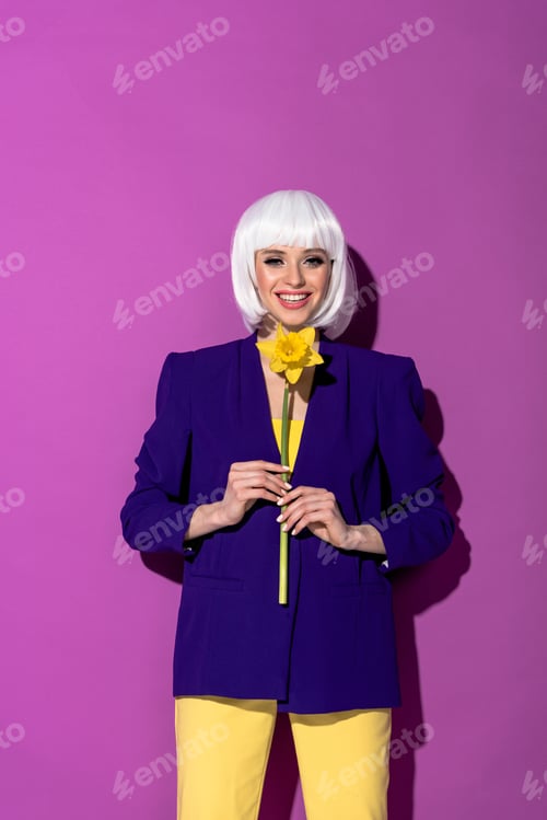 Preview: Cheerful young woman in wig holding yellow flower on purple background