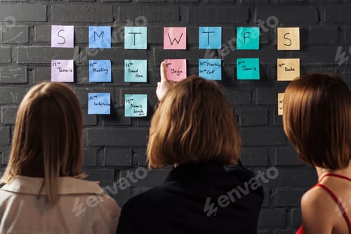 Preview: back view of three women standing near colorful sticker notes and pointing at black brick wall
