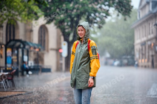 Preview: Young woman in green yellow raincoat stands happily on street under the rain