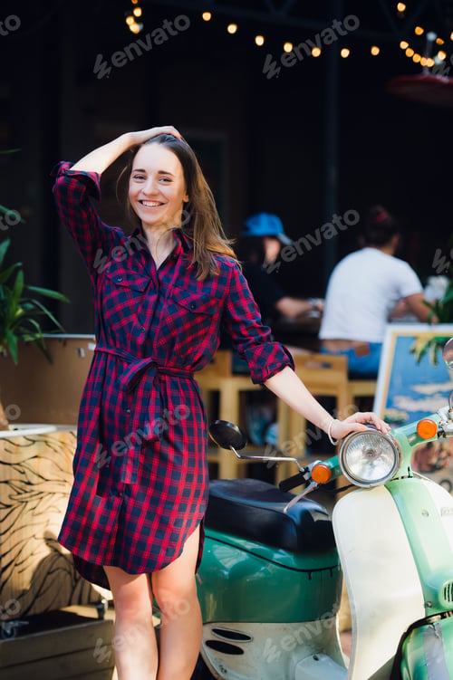 Preview: Urban fashion. Outdoor portrait of pretty young woman wearing checkered dress standing near scooter