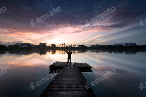 Preview: A silhouetted man standing at wooden pier during sunrise.