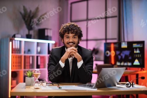 Preview: Smiling bearded man in formal suit sitting at desk with modern laptop and looking at camera