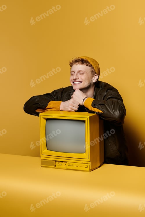 Preview: A stylish redhead man leans on a retro TV, smiling in a trendy winter outfit against yellow.