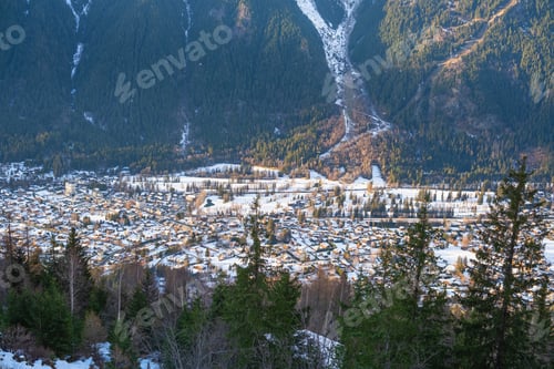 Preview: Alpine valley village covered in snow with evergreen trees in foreground and sunlit mountains