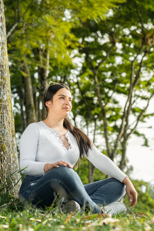 Preview: beautiful latina woman, college student sitting on the grass next to a tree observing nature