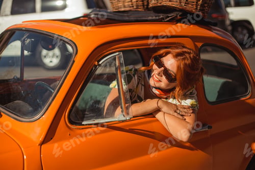 Preview: Young beautiful woman in a silk scarf and sunglasses sits behind the wheel of an orange retro car