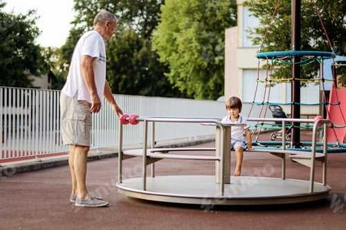 Preview: Cute happy little grandson walking with his grandfather on playground riding carousel