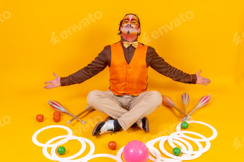 Preview: Juggler in a vest and with his face painted sitting with the juggling objects on a yellow background