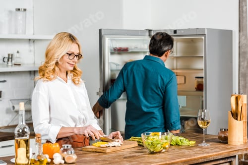 Preview: mature wife cutting vegetables and husband looking at fridge in kitchen