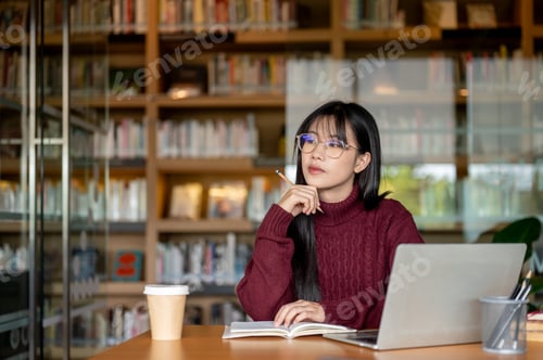 Preview: Glasses asian woman holding pencil and thinking beside laptop and coffee at wooden table in library.