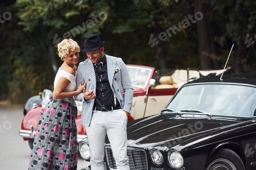 Preview: Beautiful couple in old fashioned wear near retro car with forest at background