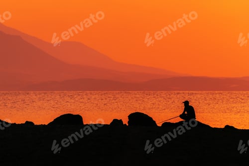 Preview: Fishing at the Aegean sea coast in summer sunrise, silhouette of fisherman with rod at the coastline