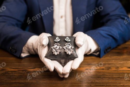 Preview: Cropped view of jewelry appraiser holding jewelry pillow with earrings at table isolated on black
