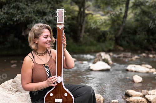 Preview: A young adult woman is smiling playing her indian tanpura sitting on a river rock
