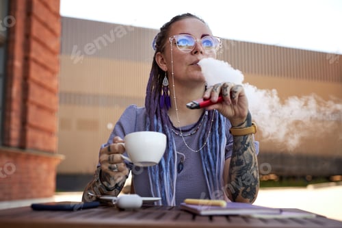 Preview: Young woman with tattoos on arms relaxing in outdoor cafe and smoking