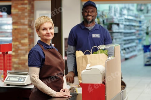 Preview: Intercultural staff of supermarket in uniform standing by checkout counter