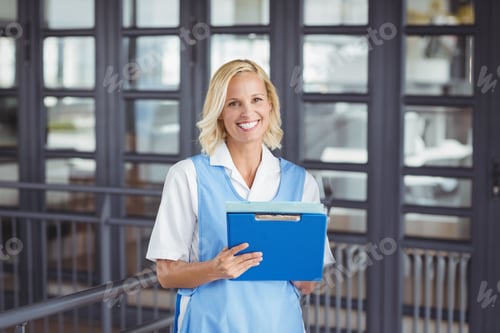 Preview: young adult woman standing in office hallway holding bright blue clipboard with both hands, smiling